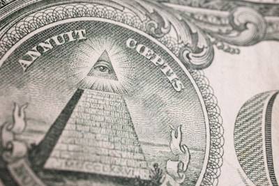 An intricate engraving of the Eye of Providence, commonly known as an all-seeing eye, is depicted within a triangular shape atop an unfinished pyramid. Surrounding the pyramid, there are Latin inscriptions and ornate scrollwork designs, suggesting symbolic and historical significance.