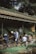 Several people are engaged in gardening work under a bamboo structure, planting and arranging plants with precision. The setting is outdoors, surrounded by trees and natural elements.