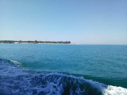 A serene ocean scene with calm blue waters extending towards the horizon. Gentle waves are visible in the foreground, while a distant island covered with lush greenery lines the horizon under a clear blue sky.