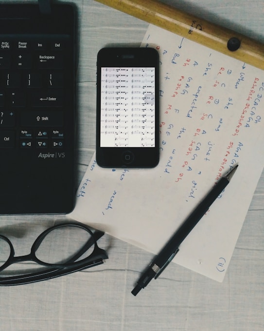 A flat lay includes a black keyboard, a smartphone displaying musical notes, handwritten notes on paper, a wooden flute, a pair of black glasses, and a black pen on a light-colored surface.