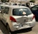 A silver compact car is shown with noticeable damage to its rear bumper and hatch, including dents and scratches. The back window has stickers, one prominently saying 'GOD IS GREAT' and another smaller one indicating 'PRINCESS ON BOARD.' The car is parked among other vehicles in an urban setting with buildings in the background.
