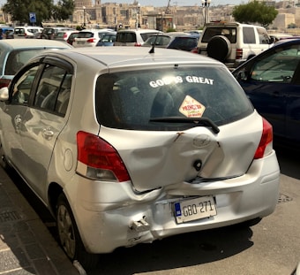 A silver compact car is shown with noticeable damage to its rear bumper and hatch, including dents and scratches. The back window has stickers, one prominently saying 'GOD IS GREAT' and another smaller one indicating 'PRINCESS ON BOARD.' The car is parked among other vehicles in an urban setting with buildings in the background.
