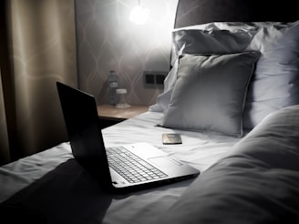A sleek laptop is open and placed on a neatly made bed alongside a smartphone. The bed has a grey pillow, and there is a nightstand with a water bottle and a glass in the background. The lighting is subtle, creating a cozy ambiance.