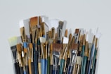 A collection of various paintbrushes with different sizes and shapes, including flat, round, and fan types. The handle colors vary, displaying shades of blue, green, and black, while the bristles range from white to brown. The arrangement is dense, showcasing the metallic bands holding the bristles in place.
