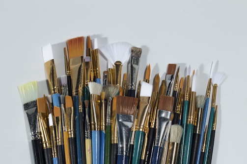A collection of various paintbrushes with different sizes and shapes, including flat, round, and fan types. The handle colors vary, displaying shades of blue, green, and black, while the bristles range from white to brown. The arrangement is dense, showcasing the metallic bands holding the bristles in place.