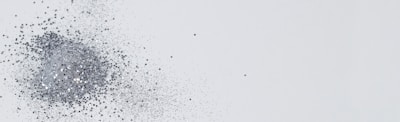A pile of fine, shiny silver glitter appears to be sprinkled across a plain, light gray background. The glitter scatters outward, fading into smaller particles toward the right side of the image, creating a dynamic and textured effect.