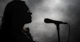 A silhouette of a person wearing glasses stands in front of a microphone against a misty, dimly lit background. The lighting creates an atmospheric scene, emphasizing the contours of the person's face and the shape of the microphone.