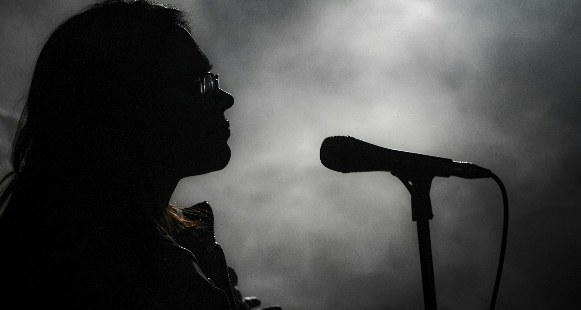 A silhouette of a person wearing glasses stands in front of a microphone against a misty, dimly lit background. The lighting creates an atmospheric scene, emphasizing the contours of the person's face and the shape of the microphone.