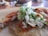 A close-up view of a taco filled with meat, chopped onions, cilantro, and a green sauce on a beige plate. The background is slightly blurred, featuring some table settings.