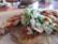 A close-up view of a taco filled with meat, chopped onions, cilantro, and a green sauce on a beige plate. The background is slightly blurred, featuring some table settings.