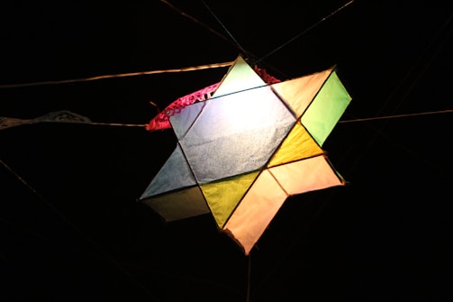 A multicolored kite with triangular and quadrilateral sections is illuminated against a dark background. The kite's panels include shades of blue, green, yellow, orange, and pink. It is tethered by strings that are visible against the night sky.