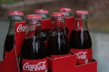 Six glass bottles of Coca-Cola in a red cardboard carrier. The bottles have metal caps with Coca-Cola branding, and they are filled with a dark liquid. The carrier features the Coca-Cola logo prominently.