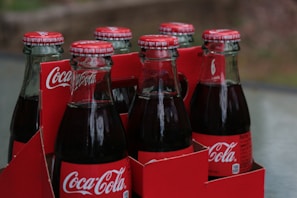 Six glass bottles of Coca-Cola in a red cardboard carrier. The bottles have metal caps with Coca-Cola branding, and they are filled with a dark liquid. The carrier features the Coca-Cola logo prominently.