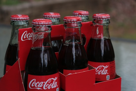 Six glass bottles of Coca-Cola in a red cardboard carrier. The bottles have metal caps with Coca-Cola branding, and they are filled with a dark liquid. The carrier features the Coca-Cola logo prominently.