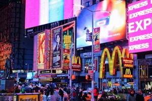 A vibrant city street scene featuring bright neon lights and large advertisements for theatrical productions and fast food. People are walking along the street, surrounded by illuminated signs, including those for McDonald's and various Broadway shows. The scene is bustling with activity, capturing the lively atmosphere of a busy urban area.