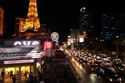A vibrant night scene of the Las Vegas Strip, filled with illuminated buildings, bustling streets, and heavy traffic. Iconic neon signs glow prominently, and the replica of the Eiffel Tower stands out on the left side, brilliantly lit. Bright storefronts and numerous cars contribute to the lively atmosphere.