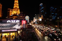 A vibrant night scene of the Las Vegas Strip, filled with illuminated buildings, bustling streets, and heavy traffic. Iconic neon signs glow prominently, and the replica of the Eiffel Tower stands out on the left side, brilliantly lit. Bright storefronts and numerous cars contribute to the lively atmosphere.