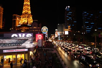 A vibrant night scene of the Las Vegas Strip, filled with illuminated buildings, bustling streets, and heavy traffic. Iconic neon signs glow prominently, and the replica of the Eiffel Tower stands out on the left side, brilliantly lit. Bright storefronts and numerous cars contribute to the lively atmosphere.