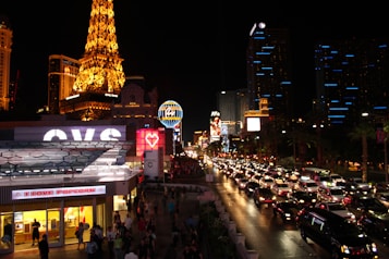 A vibrant night scene of the Las Vegas Strip, filled with illuminated buildings, bustling streets, and heavy traffic. Iconic neon signs glow prominently, and the replica of the Eiffel Tower stands out on the left side, brilliantly lit. Bright storefronts and numerous cars contribute to the lively atmosphere.