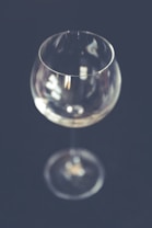 A clear, empty wine glass is centered against a dark, blurred background. The glass reflects subtle light, highlighting its smooth contours and elegant shape.