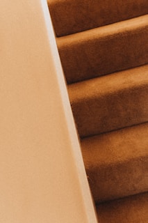 A close-up view of a carpeted staircase with beige walls. The carpeting is a warm brown color, and the perspective is angled, focusing on the texture and the repeating pattern of the steps.