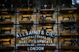 Rows of vintage Singer sewing machines are displayed on wooden shelves against a rustic brick wall. Below the sewing machines, there's white text painted directly onto the wall, featuring the brand name 'AllSaints Co. Ltd.', along with a tagline about manufacturing quality garments in Spitalfields, London.