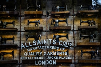 Rows of vintage Singer sewing machines are displayed on wooden shelves against a rustic brick wall. Below the sewing machines, there's white text painted directly onto the wall, featuring the brand name 'AllSaints Co. Ltd.', along with a tagline about manufacturing quality garments in Spitalfields, London.