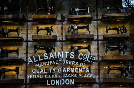 Rows of vintage Singer sewing machines are displayed on wooden shelves against a rustic brick wall. Below the sewing machines, there's white text painted directly onto the wall, featuring the brand name 'AllSaints Co. Ltd.', along with a tagline about manufacturing quality garments in Spitalfields, London.