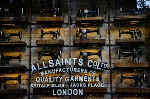 Rows of vintage Singer sewing machines are displayed on wooden shelves against a rustic brick wall. Below the sewing machines, there's white text painted directly onto the wall, featuring the brand name 'AllSaints Co. Ltd.', along with a tagline about manufacturing quality garments in Spitalfields, London.