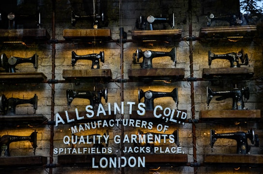 Rows of vintage Singer sewing machines are displayed on wooden shelves against a rustic brick wall. Below the sewing machines, there's white text painted directly onto the wall, featuring the brand name 'AllSaints Co. Ltd.', along with a tagline about manufacturing quality garments in Spitalfields, London.