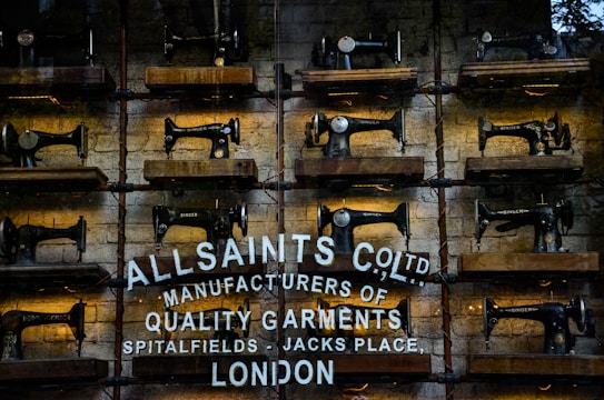 Rows of vintage Singer sewing machines are displayed on wooden shelves against a rustic brick wall. Below the sewing machines, there's white text painted directly onto the wall, featuring the brand name 'AllSaints Co. Ltd.', along with a tagline about manufacturing quality garments in Spitalfields, London.