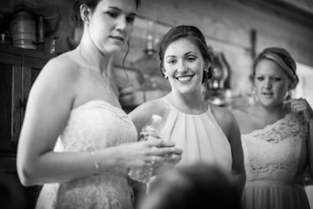 A group of three young women, dressed in elegant attire, stand indoors in a warmly lit setting. One woman, smiling brightly, holds a clear water bottle, while the others look on with relaxed expressions. The background features wooden cabinets and various decorative items.