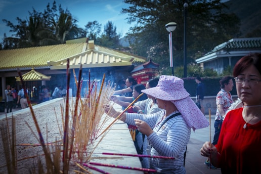 A group of people are engaged in a ritual involving incense sticks at a traditional outdoor setting. The foreground features a woman wearing a wide-brimmed floral hat, lighting and placing incense sticks into a sand-filled altar. The background shows additional participants and a building with yellow-tiled roofs, surrounded by trees under a clear sky.