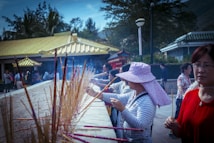 A group of people are engaged in a ritual involving incense sticks at a traditional outdoor setting. The foreground features a woman wearing a wide-brimmed floral hat, lighting and placing incense sticks into a sand-filled altar. The background shows additional participants and a building with yellow-tiled roofs, surrounded by trees under a clear sky.