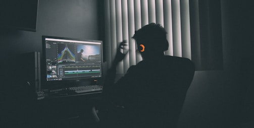 A person sitting at a computer desk wearing headphones, surrounded by dim lighting. The computer screen displays video editing software with various panels showing timelines and video footage. Blinds in the background allow minimal light into the room, creating a silhouette of the person.