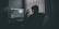 A person sitting at a computer desk wearing headphones, surrounded by dim lighting. The computer screen displays video editing software with various panels showing timelines and video footage. Blinds in the background allow minimal light into the room, creating a silhouette of the person.