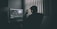 A person sitting at a computer desk wearing headphones, surrounded by dim lighting. The computer screen displays video editing software with various panels showing timelines and video footage. Blinds in the background allow minimal light into the room, creating a silhouette of the person.