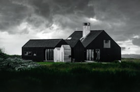 A modern, dark-colored house with multiple pitched roofs stands in a lush green landscape under a dramatic, cloudy sky. The architecture contrasts sharply with the natural surroundings, creating a striking visual. The sky is overcast, hinting at an impending storm.