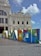A white, ornate historic building with classical architectural features such as columns and decorative sculptures is prominently displayed. In front of the building, a colorful structure spelling out 'SALVADOR' is positioned, featuring vivid, abstract designs and the Rio 2016 Olympics logo. The sky is clear with some scattered clouds.
