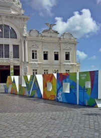 A white, ornate historic building with classical architectural features such as columns and decorative sculptures is prominently displayed. In front of the building, a colorful structure spelling out 'SALVADOR' is positioned, featuring vivid, abstract designs and the Rio 2016 Olympics logo. The sky is clear with some scattered clouds.