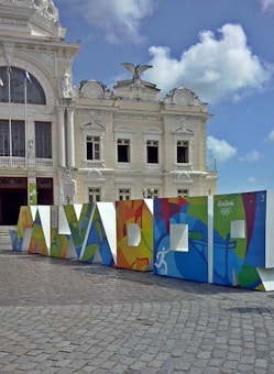 A white, ornate historic building with classical architectural features such as columns and decorative sculptures is prominently displayed. In front of the building, a colorful structure spelling out 'SALVADOR' is positioned, featuring vivid, abstract designs and the Rio 2016 Olympics logo. The sky is clear with some scattered clouds.