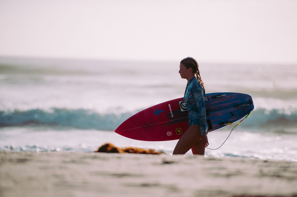 Donna che trasporta tavola da surf rossa e blu che cammina sulla riva durante il giorno