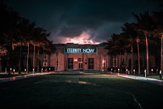 A building is illuminated by neon lights against a dramatic, cloudy twilight sky. The phrase 'ETERNITY NOW' glows in blue above the entrance. Palm trees line the pathway leading to the structure, while small lights cast a warm glow along the walkway.