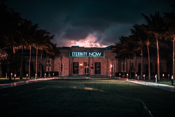A building is illuminated by neon lights against a dramatic, cloudy twilight sky. The phrase 'ETERNITY NOW' glows in blue above the entrance. Palm trees line the pathway leading to the structure, while small lights cast a warm glow along the walkway.