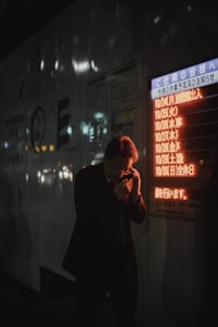 A person in a dark suit is holding a phone to their ear, standing near a bright, illuminated board displaying text in Japanese. The setting appears to be urban and dimly lit, suggesting it is nighttime. The orange and red glow from the board contrasts with the dark surroundings.