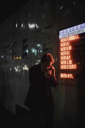 A person in a dark suit is holding a phone to their ear, standing near a bright, illuminated board displaying text in Japanese. The setting appears to be urban and dimly lit, suggesting it is nighttime. The orange and red glow from the board contrasts with the dark surroundings.