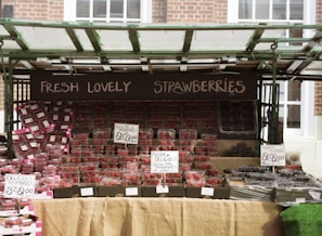 A market stall displays a variety of fresh berries, primarily strawberries, along with raspberries and blueberries. The strawberries are prominently arranged in clear plastic containers, neatly stacked in rows. Signs with handwritten text highlight the prices and emphasize the freshness and origin of the berries. The stall is sheltered by a green-framed canopy.