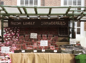A market stall displays a variety of fresh berries, primarily strawberries, along with raspberries and blueberries. The strawberries are prominently arranged in clear plastic containers, neatly stacked in rows. Signs with handwritten text highlight the prices and emphasize the freshness and origin of the berries. The stall is sheltered by a green-framed canopy.