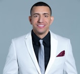 A person is wearing a white suit jacket with a black shirt and a light gray tie. A maroon pocket square is tucked into the breast pocket. The background is a solid light gray color.