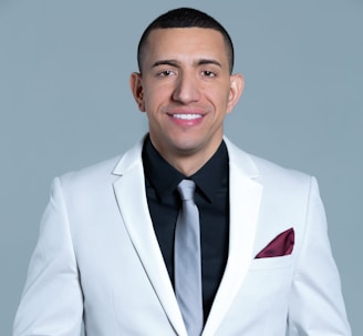 A person is wearing a white suit jacket with a black shirt and a light gray tie. A maroon pocket square is tucked into the breast pocket. The background is a solid light gray color.